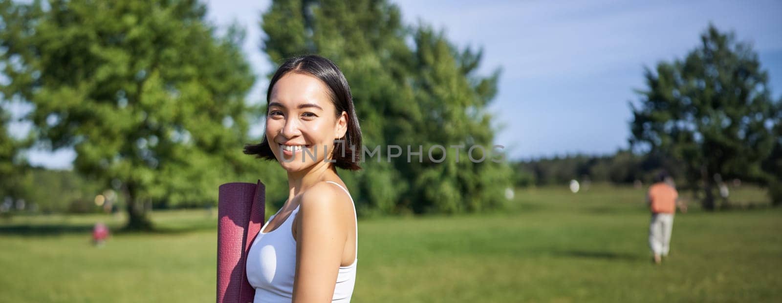 Smiling fitness girl with rubber mat, stands in park wearing uniform for workout and sport activities, does yoga outdoors on lawn.