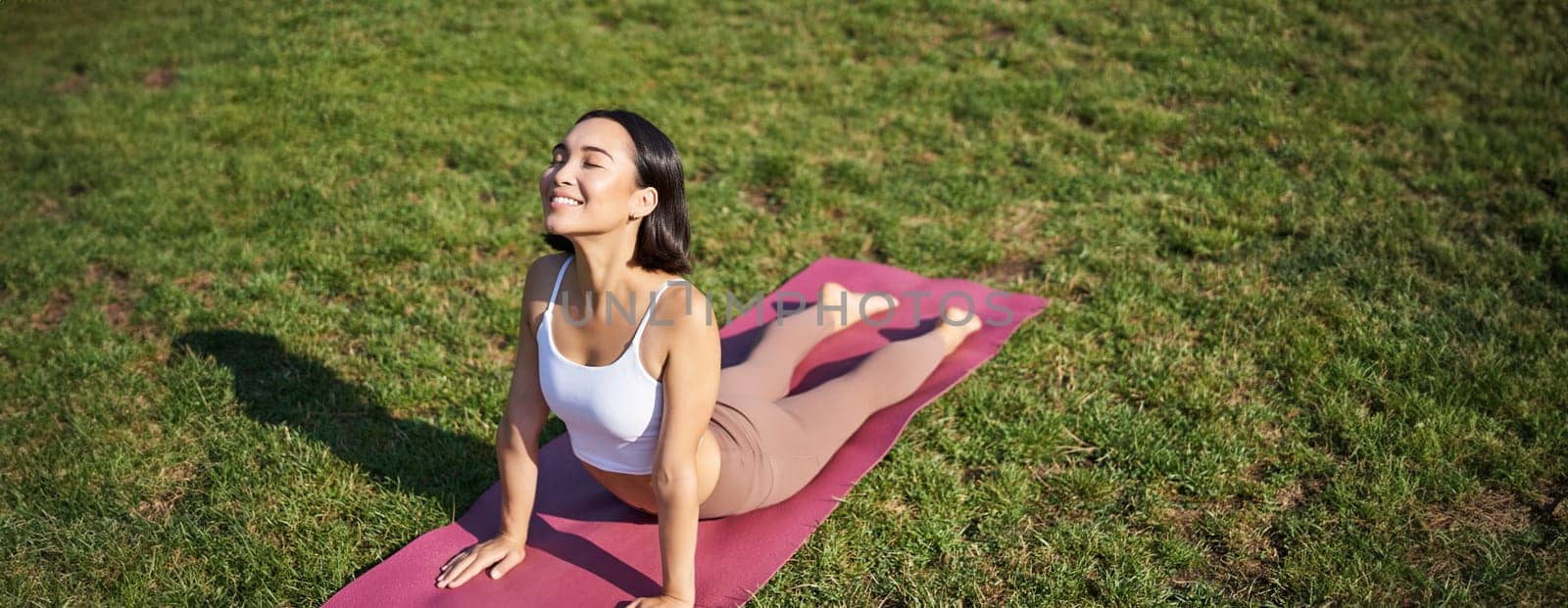 Smiling young sportswoman stretches on rubber mat in park, does yoga asana exercises, workout on fresh air in fitness clothing by Benzoix