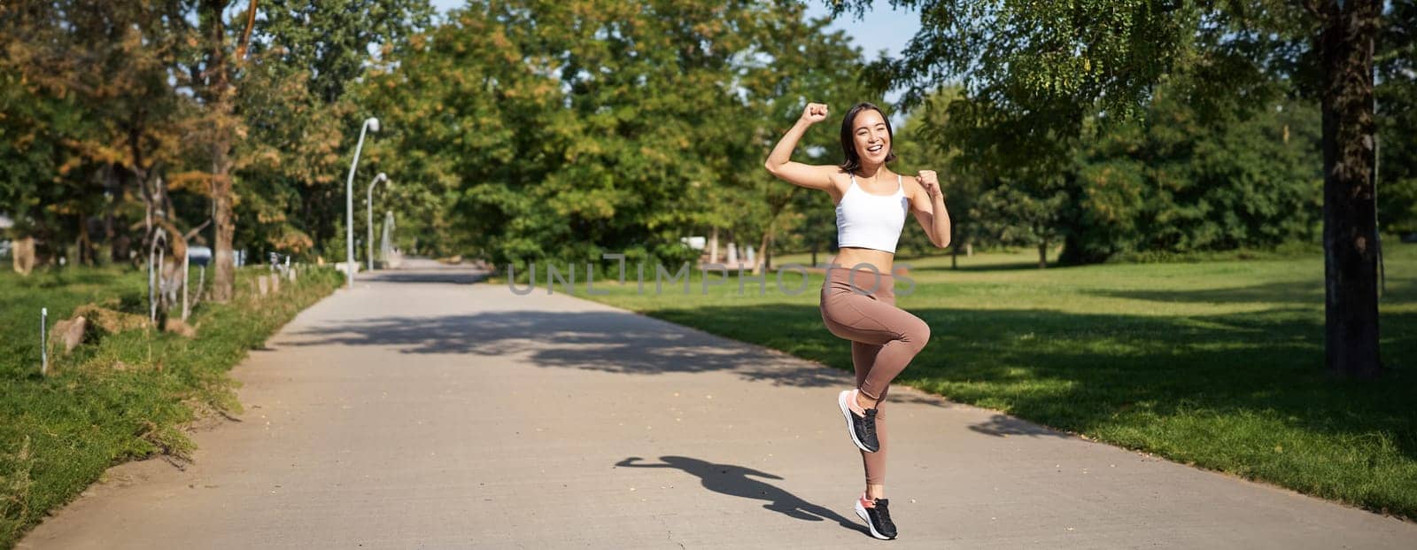 Excited young asian woman winning, finish running in park, saying yes, lifting hand up in triumph, celebrating victory or success.