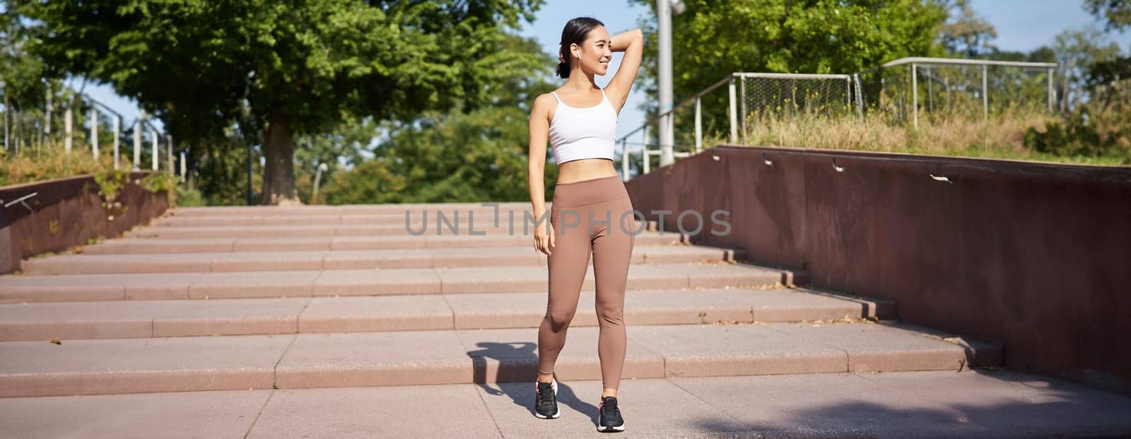 Sportswoman jogging outdoors. Smiling asian fitness girl in wireless earphones, listening music and running on road, workout.