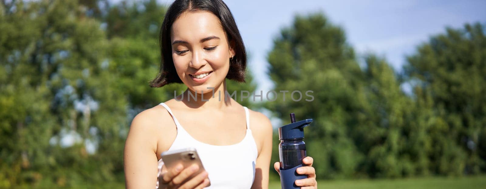 Smiling fitness girl drinks water, checks her app on smartphone and looks happy, stays hydrated on fresh air, sunny day in park.