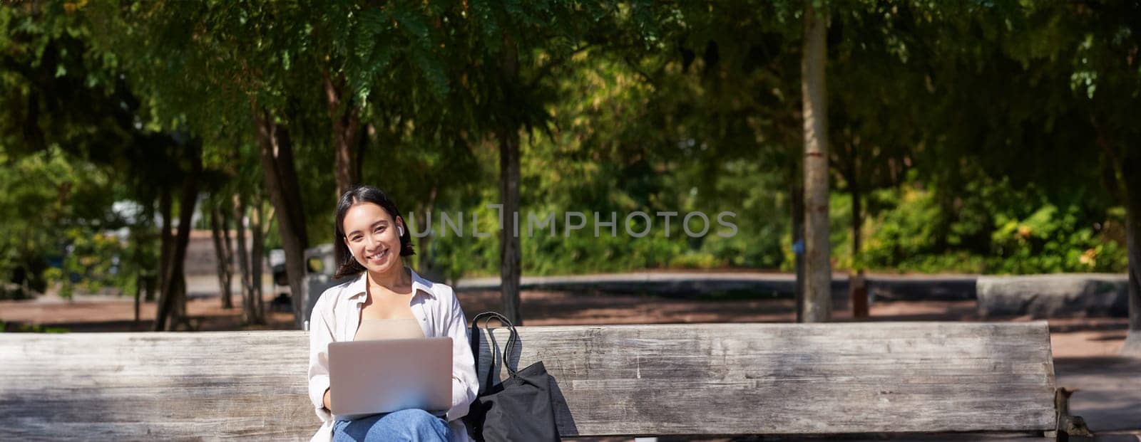 Portrait of asian woman sitting with laptop on bench in park, listening music with wireless headphones, doing homework, working on remote by Benzoix