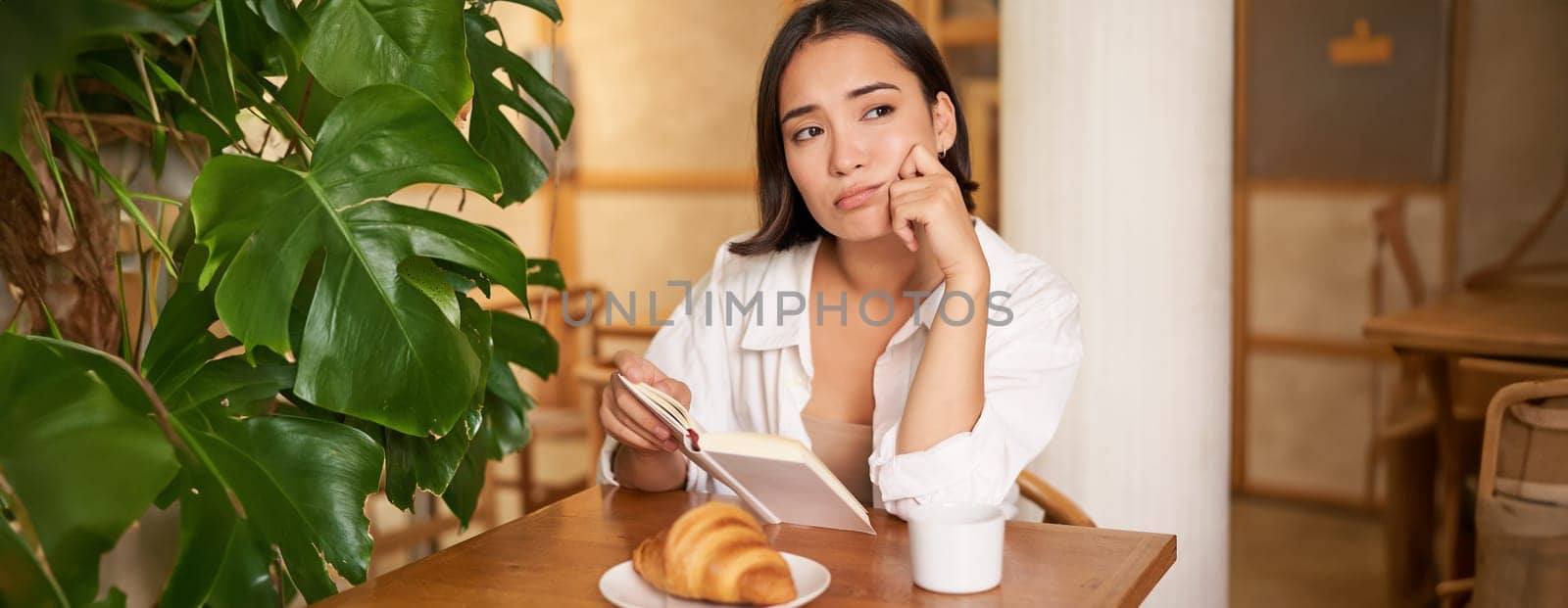 Young woman feels uneasy, reads sad book and sits in cafe with cup of coffee, looks around with sulking unhappy face.