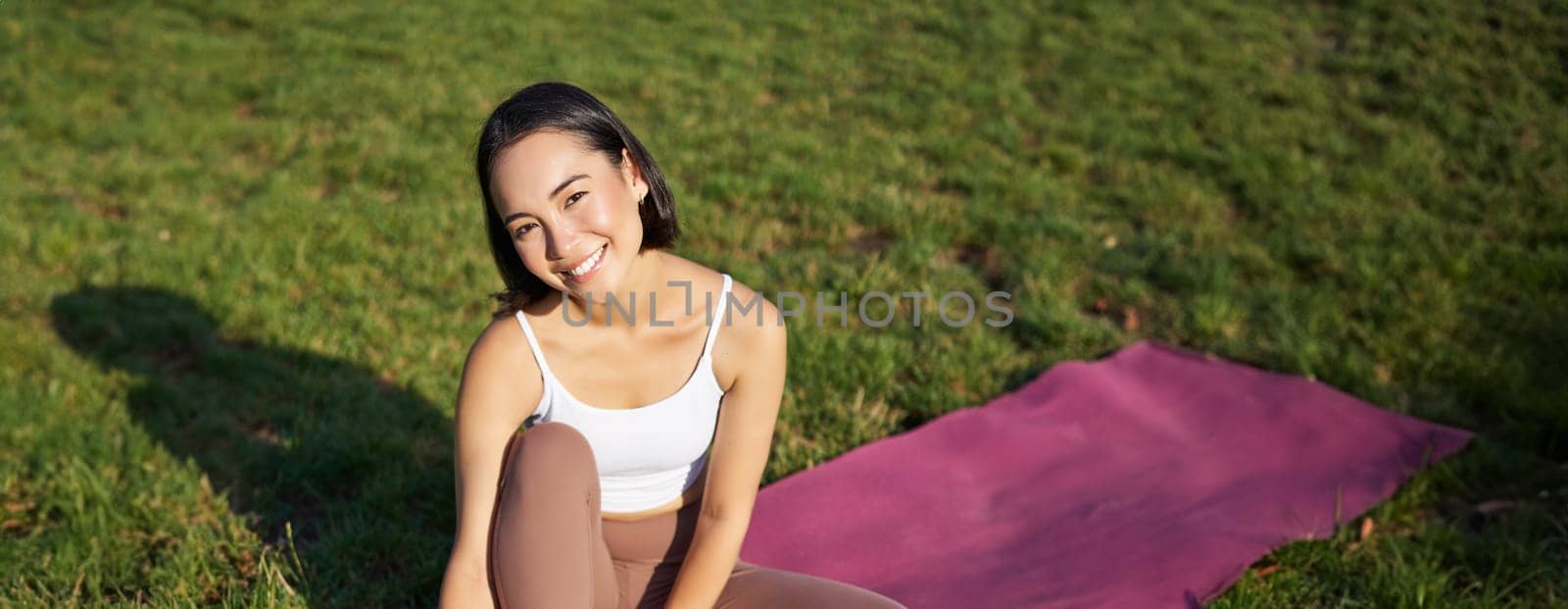 Vertical shot of smiling asian girl meditating, sitting on yoga mat with smartphone, doing yoga on fresh air in park, watching online fitness instructor by Benzoix