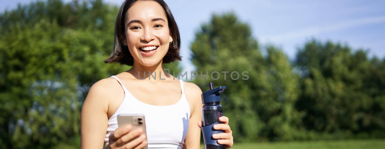 Sportswoman workout with smartphone, drinks water, holds mobile phone and smiles at camera, stands in park.
