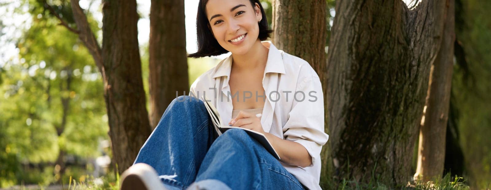 Portrait of happy asian girl sitting in park and writing in her diary. Young woman doing homework on fresh air, sitting near tree and smiling by Benzoix