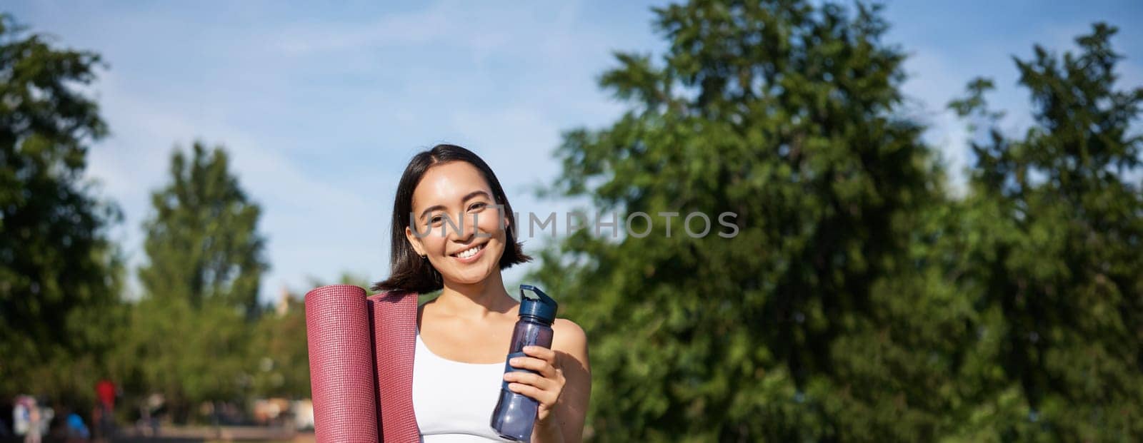 Portrait of young slim and healthy korean girl doing workout in park, standing with water bottle and rubber mat for execises on green lawn, smiling happily by Benzoix