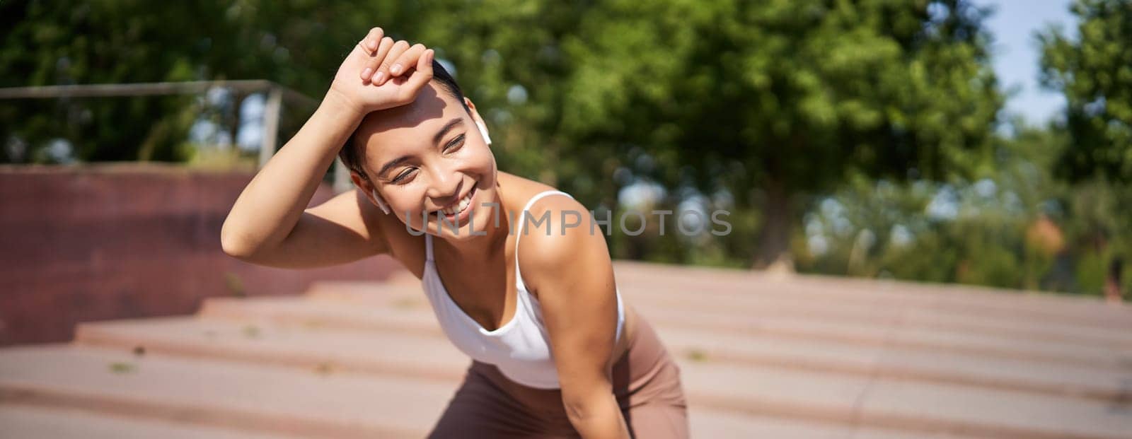Portrait of asian woman taking break, breathing heavily and panting after running, jogger standing and wiping sweat off forehead, smiling pleased by Benzoix