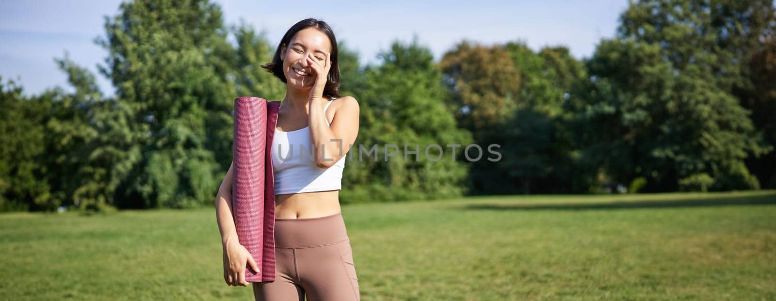 Portrait of asian girl smiles and laughs, stands with sports equipment, rubber mat, wears uniform for workout and exercises in park by Benzoix