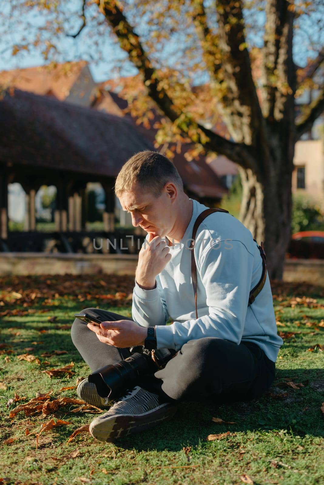 Professional Photographer Taking Picture Of Beautiful Autumn Park. Man Professional Photographer Sit With Camera And With Smartphone In Autumn Park. Retouched, Vibrant Colors, Brownish Tones. by Andrii_Ko