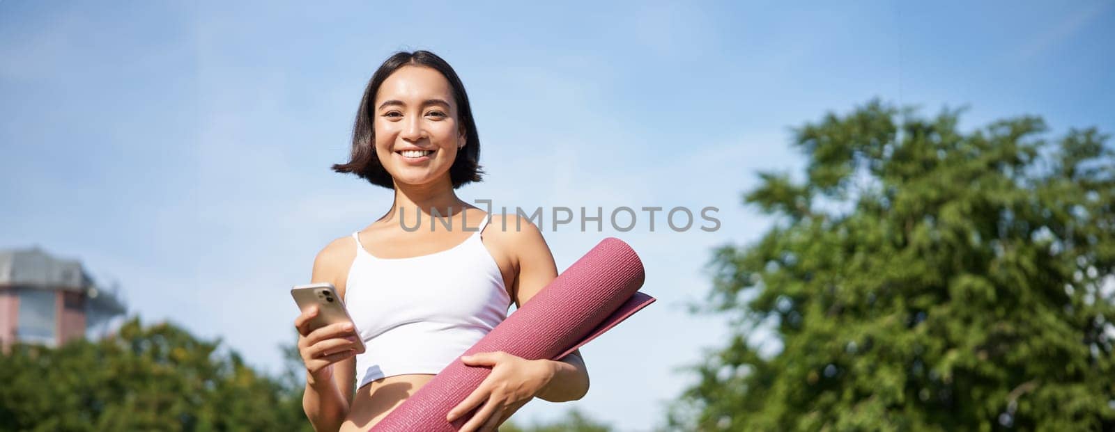 Portrait of young asian woman checking her phone during workout, walking in park with rubber mat, going to the gym, holding smartphone.