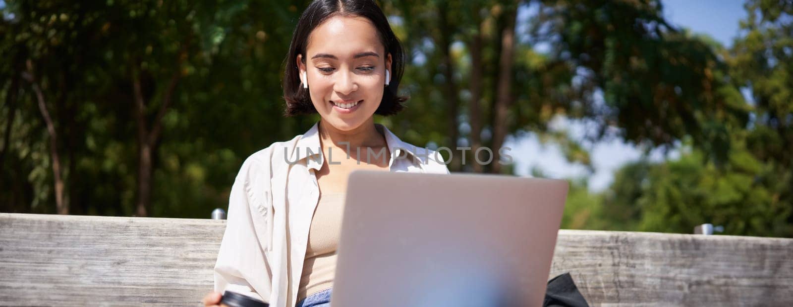 Asian girl sitting with laptop and wireless earphones, drinking coffee, looking at screen, doing homework, working on remote.