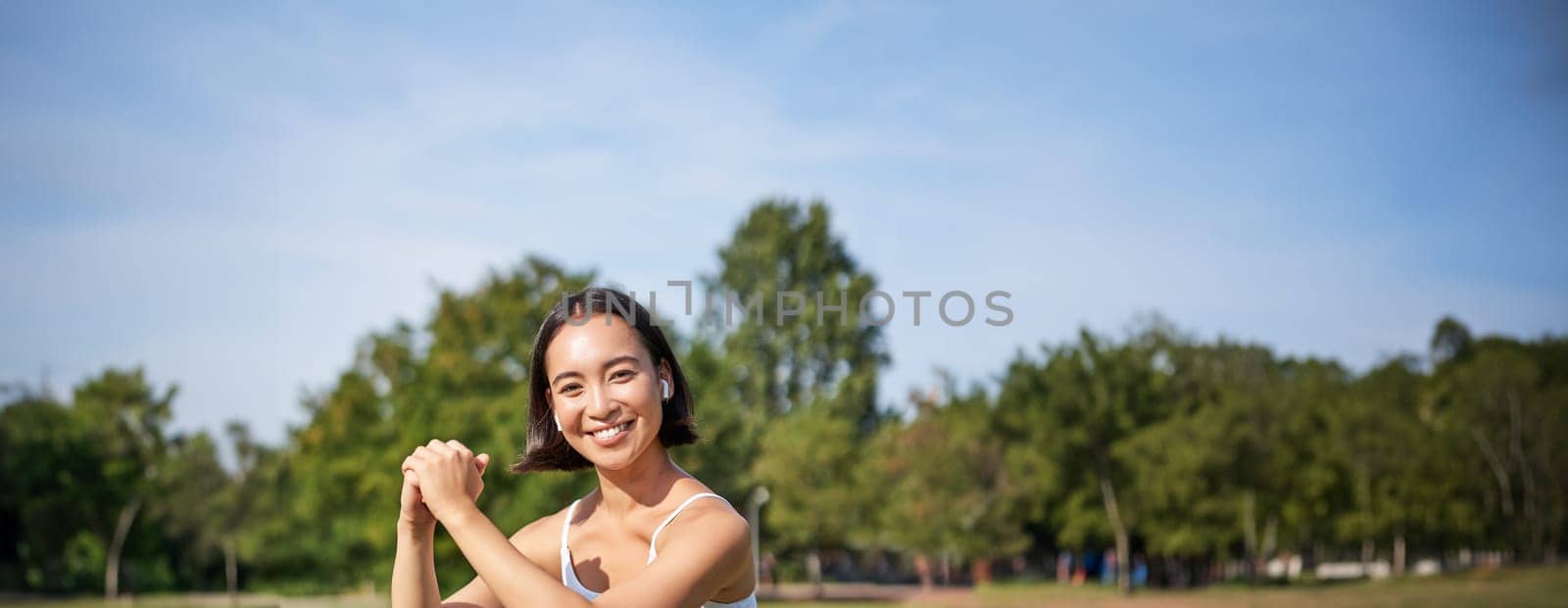 Vertical shot of young fit woman does squats in park, using stretching band on legs, smiling pleased while workout.