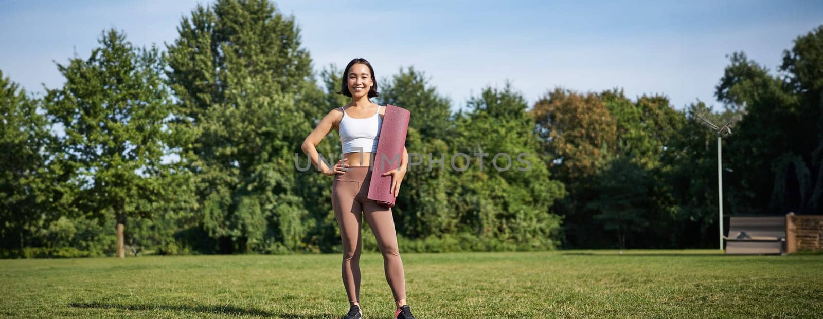 Confident and sporty young asian woman, standing in park with rubber mat, wearing sportswoman, smiling at camera, workout on fresh air by Benzoix