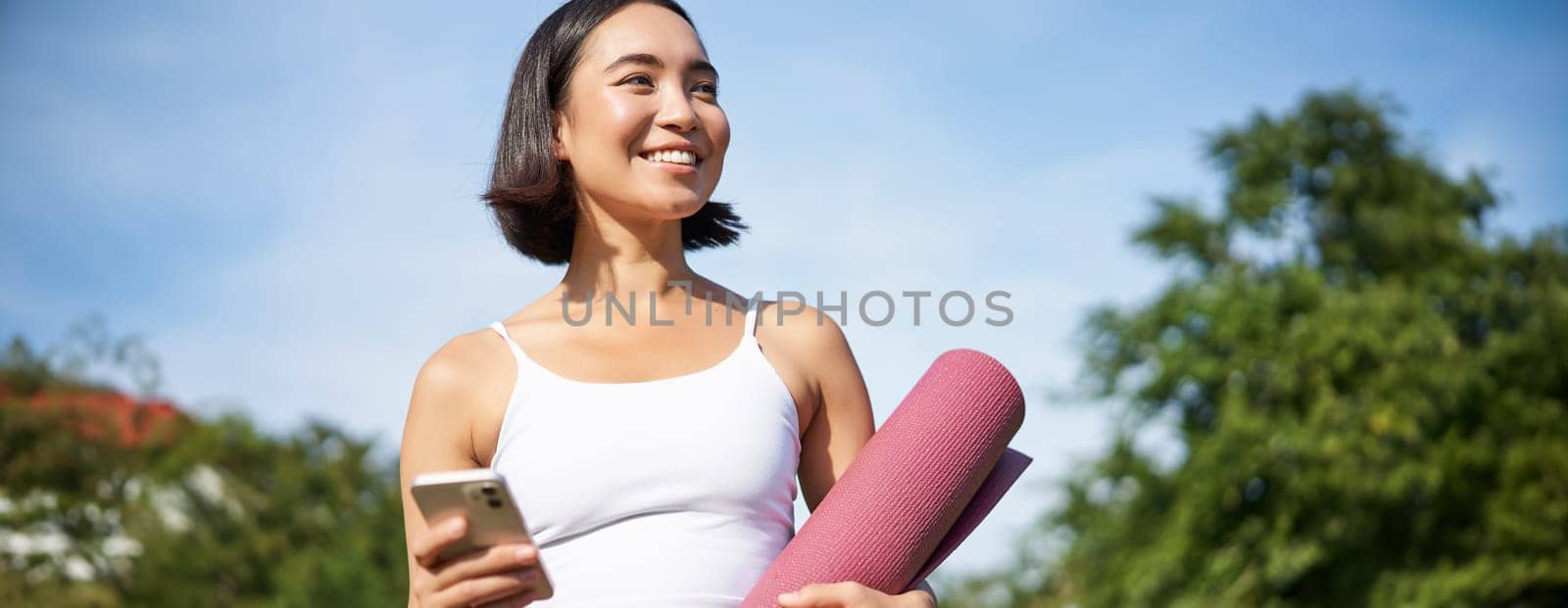 Portrait of young asian woman checking her phone during workout, walking in park with rubber mat, going to the gym, holding smartphone by Benzoix