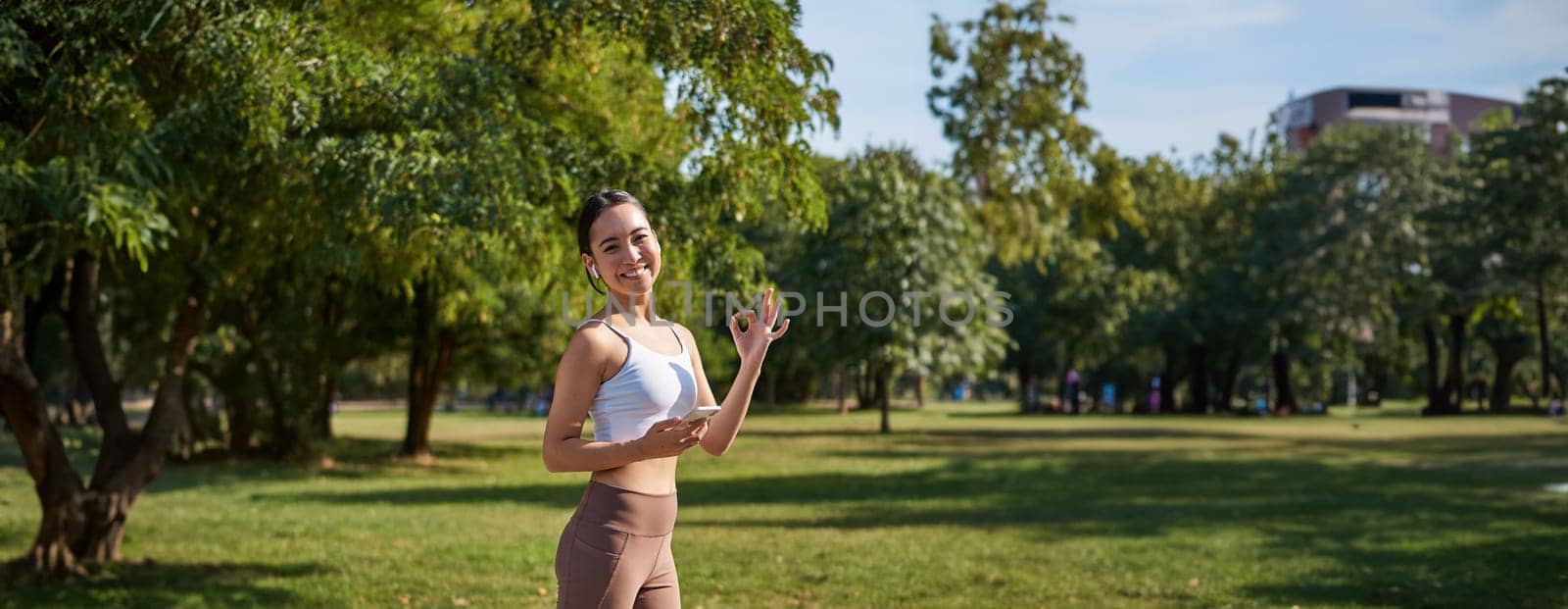 Sport and wellbeing. Young sportswoman in park, listen music and smiling, workout outdoors, jogging on streets of city.