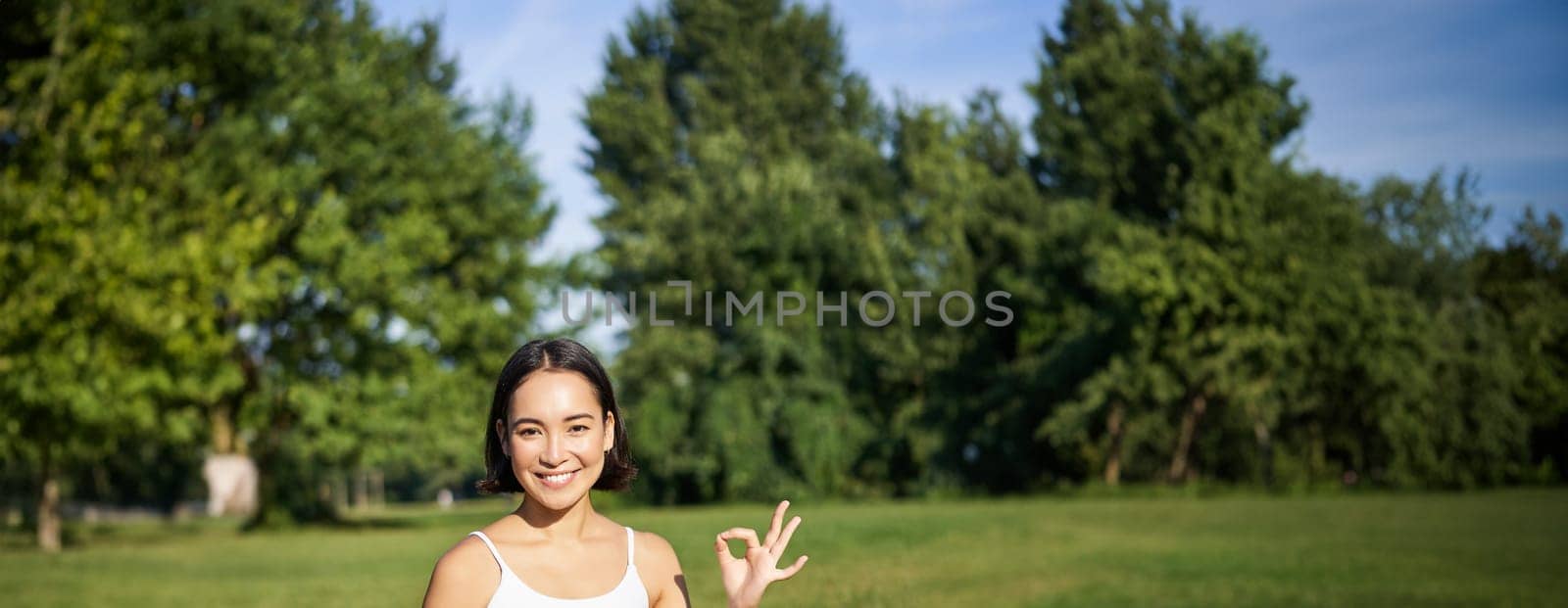 Vertical shot of asian woman shows okay sign, recommending yoga training online, meditation app, doing exercises on fresh air in park by Benzoix