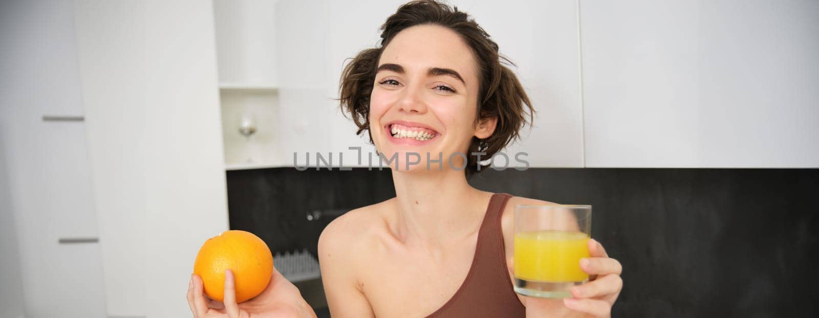 Image of sportswoman, fitness girl holding glass of juice and an orange, smiling, drinking vitamin beverage after workout, standing in her kitchen at home. Healthy lifestyle and sport concept.