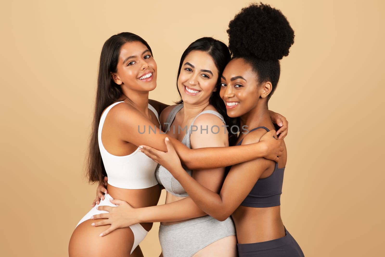Three radiant women from diverse backgrounds share moment of joy and unity, embracing their natural beauty and confidence in underwear against beige backdrop