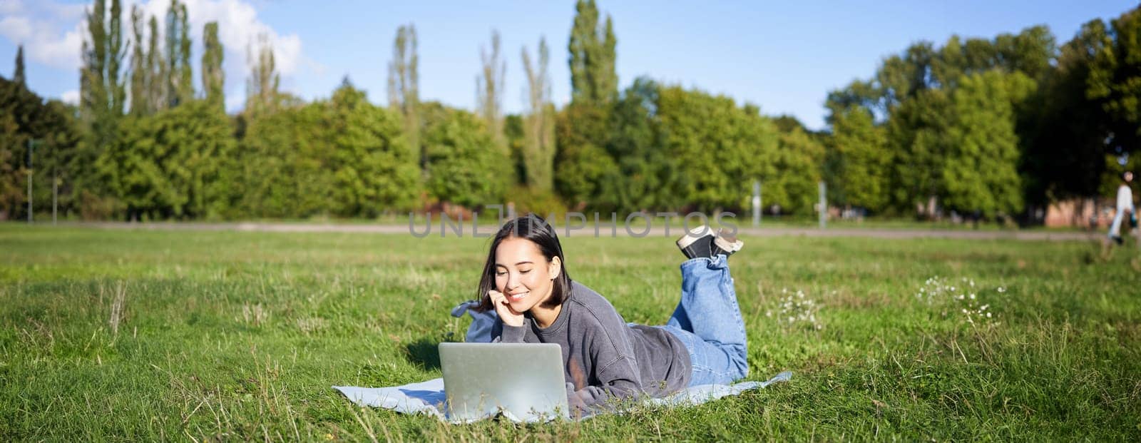 Portrait of young happy asian girl lying on blanket in park, watching videos and browsing internet on her laptop by Benzoix