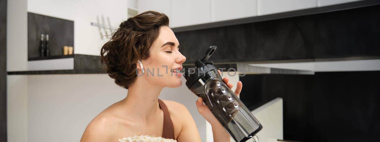 Smiling active young woman in sportswear, sits on yoga mat with water bottle, drinks stays hydrated during workout training session from home, fitness in the house on floor.