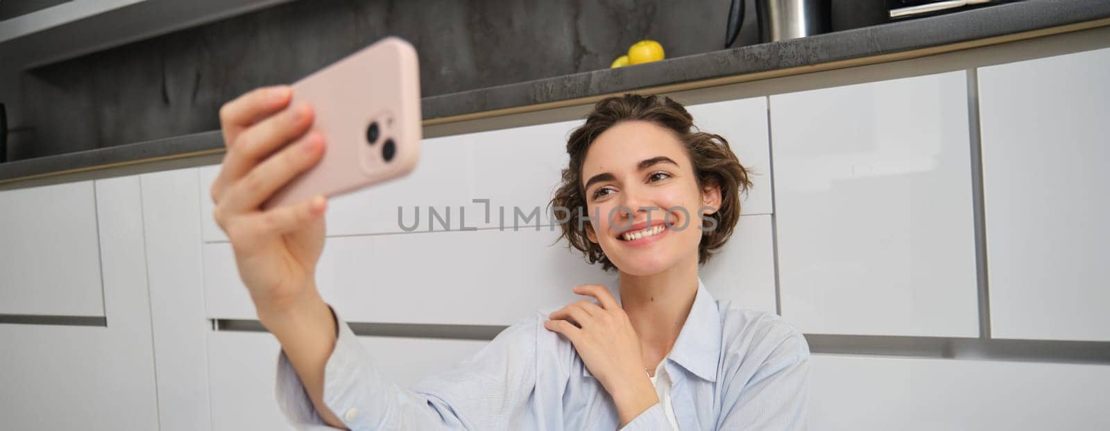 Portrait of young woman sits on kitchen floor with telephone, takes selfie on smartphone with app filters, poses for photo on mobile phone.