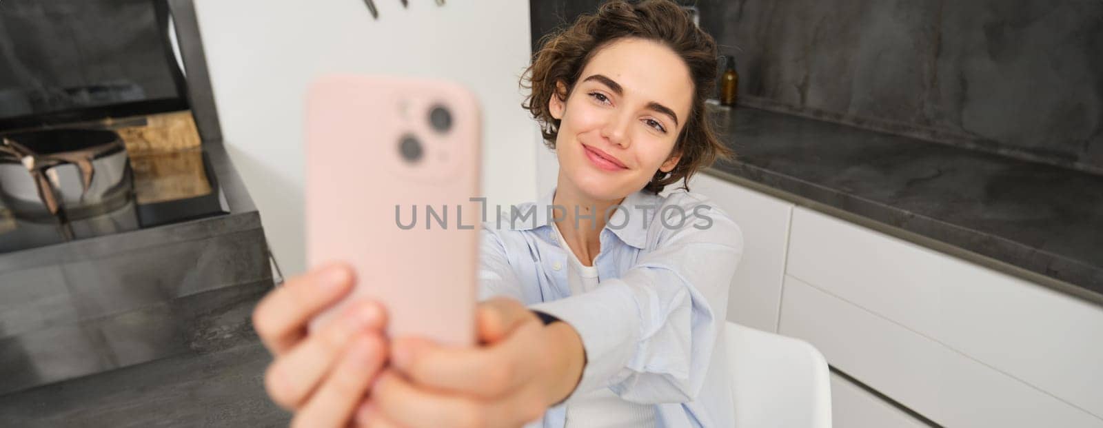 Beautiful girl takes selfie on smartphone in her kitchen, makes photos at home on mobile phone.