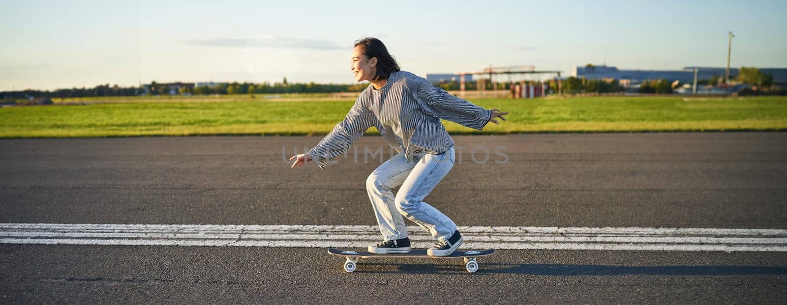 Happy skater girl riding her skateboard and having fun on empty street. Smiling woman enjoying cruiser ride on sunny road by Benzoix