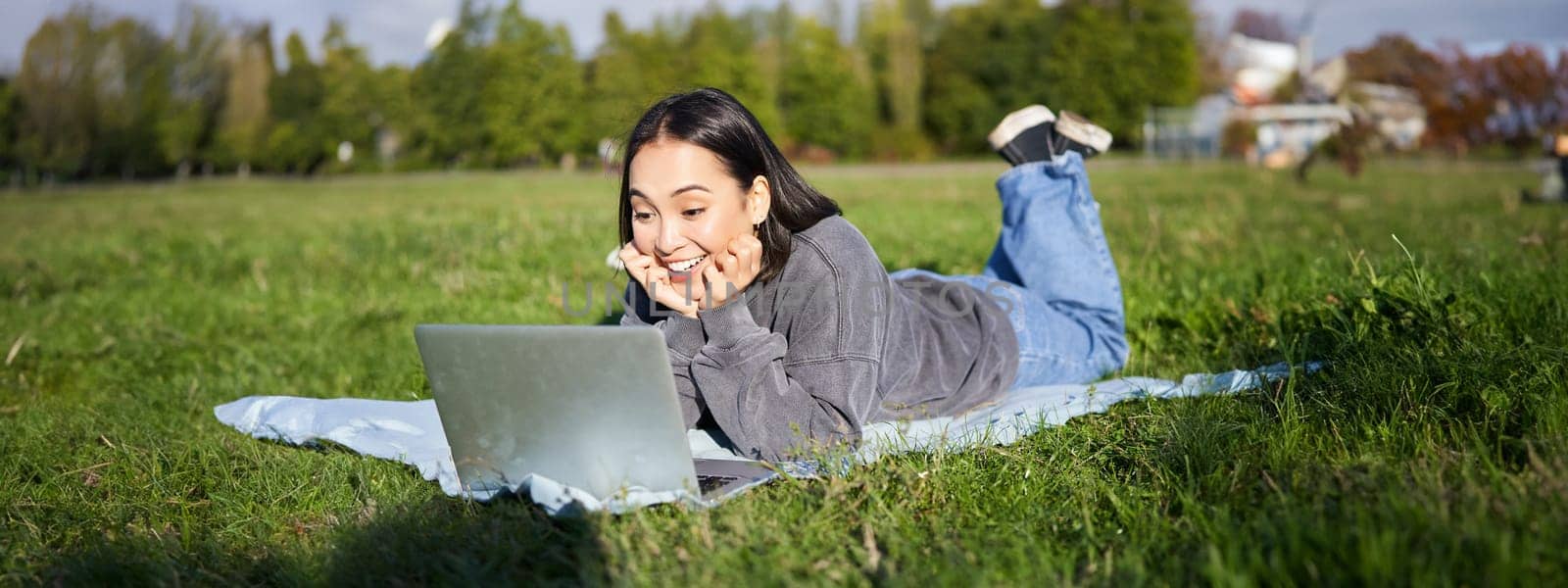 Portrait of beautiful girl lying in park and looking with surprised face at her laptop, watching videos, chatting with friends while relaxing outdoors by Benzoix