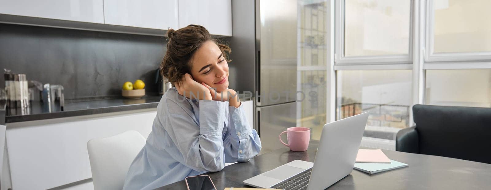 Portrait of beautiful brunette girl, contemplating smth on computer, watching video on laptop and smiling, looking dreamy at screen, sitting at home in kitchen with paperwork, working on remote.
