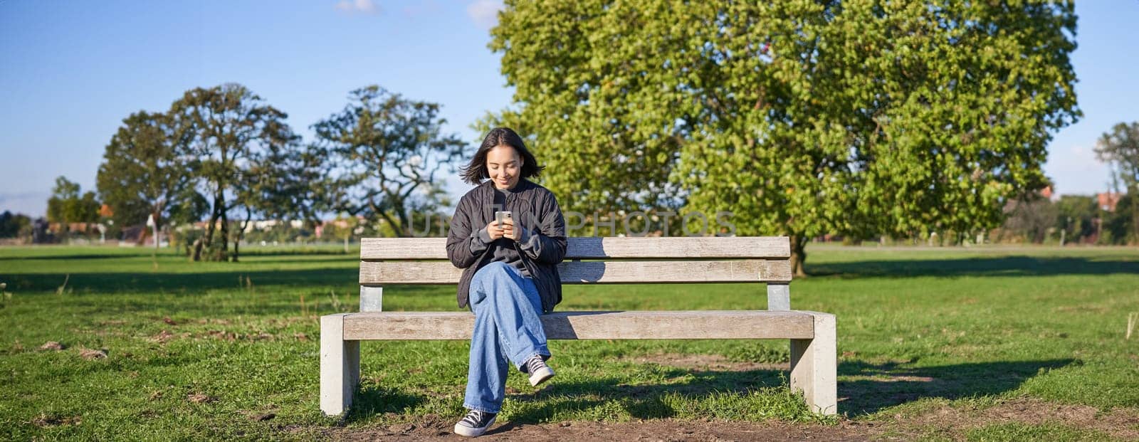 Young woman sitting alone on bench in park, using mobile phone, looking at screen, texting, sending messages in app chat.