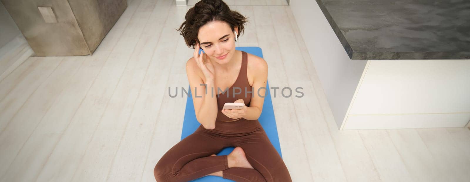 Portrait of fitness girl listens music, workout from home on yoga mat, uses smartphone sport app, watches training session, online exercises.