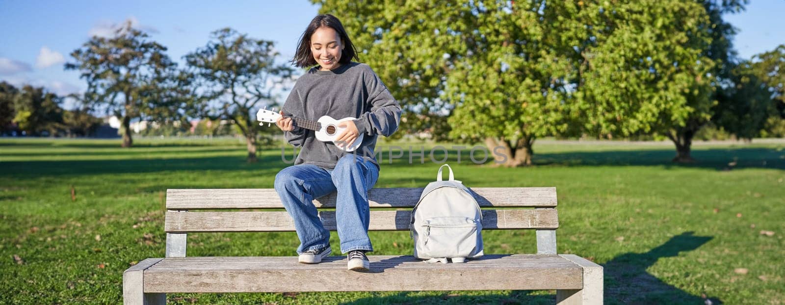Portrait of young hipster girl sits on bench and plays ukulele, sings along, relaxing in park with her musical instrument.