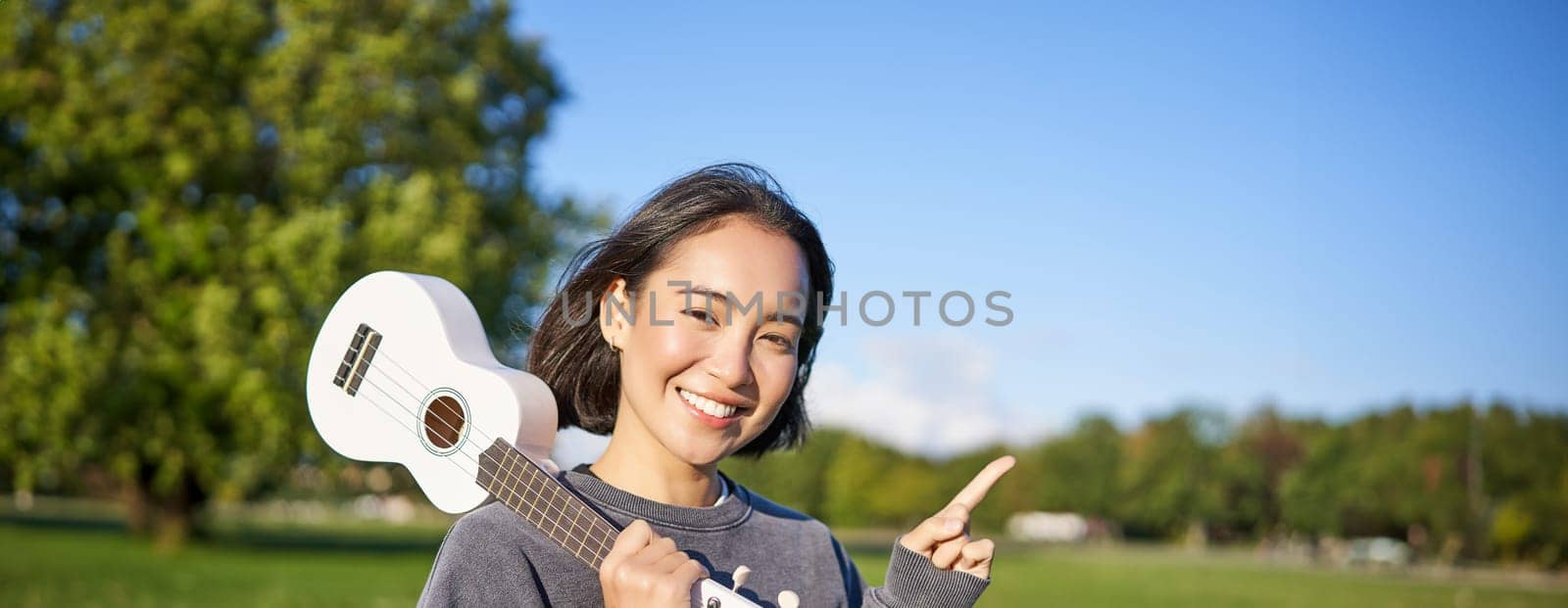 Portrait of asian smiling girl, holding ukulele over shoulder, pointing finger at copy space, banner or logo.