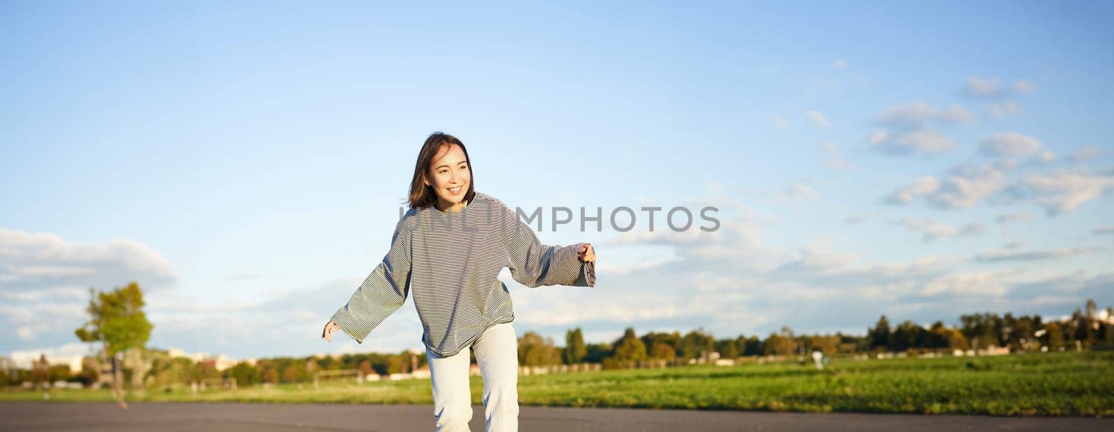 Cute asian girl riding skateboard, skating on road and smiling. Skater on cruiser longboard enjoying outdoors on sunny day.