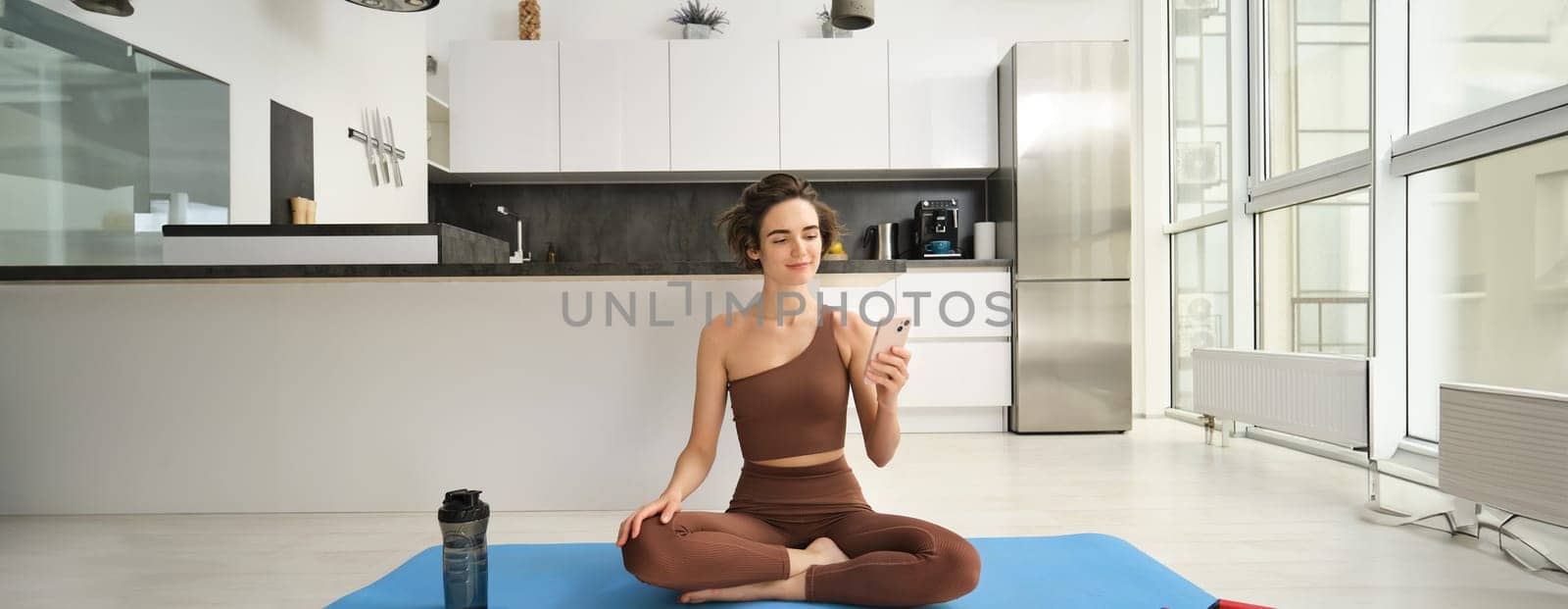 Sport app and wellbeing. Young woman, athlete sitting on floor at home with smartphone, doing yoga, using workout app on mobile phone, doing exercises at home. Copy space
