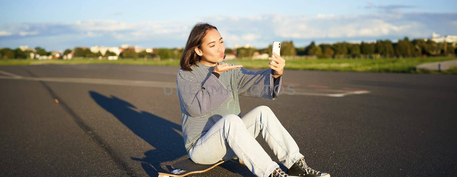 Happy asian girl sits on skateboard, takes selfie with longboard, makes cute faces, sunny day outdoors by Benzoix