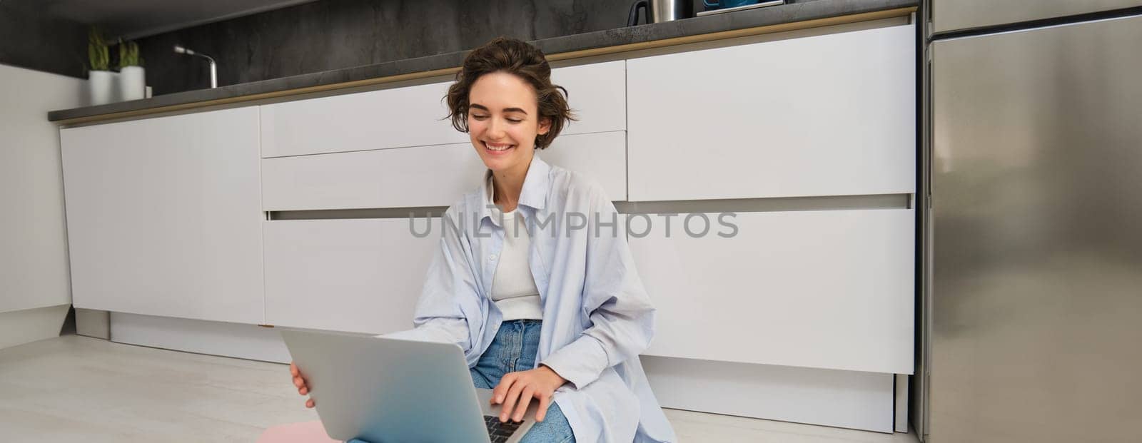 Portrait of young woman using laptop, sitting on kitchen floor, doing her homework, online chatting with friends on computer.