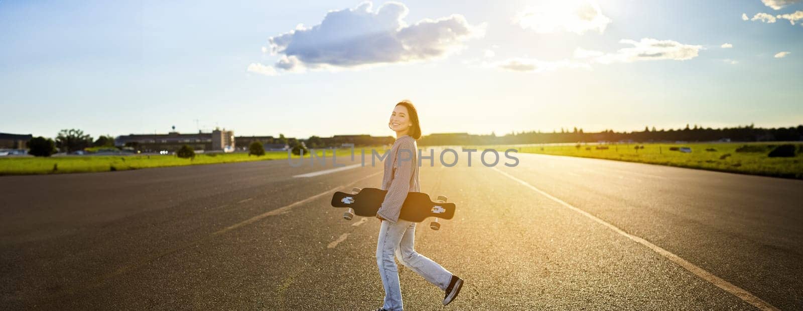 Asian girl with skateboard standing on road during sunset. Skater posing with her long board, cruiser deck during training.