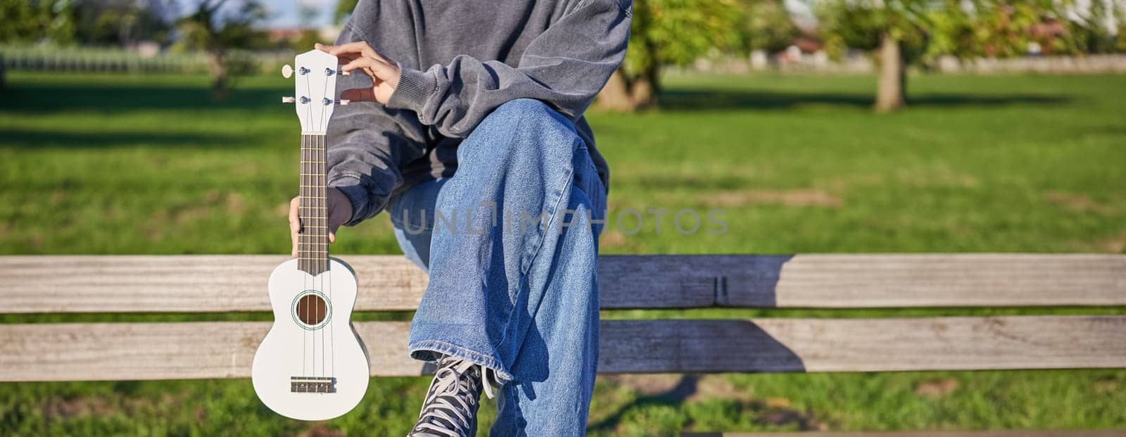 Cropped shot of young girl in sneakers and jeans, hands holding ukulele musical instrument while she sits on bench in green sunny park.