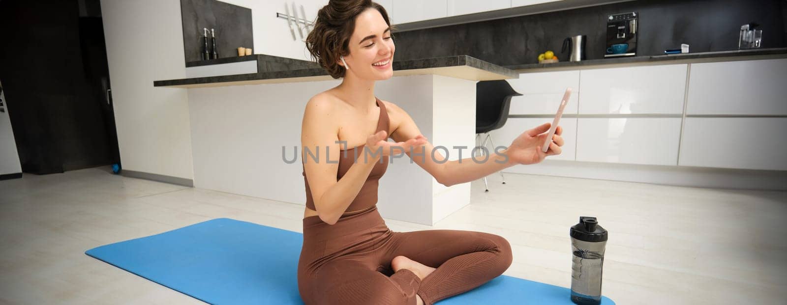 Portrait of happy smiling, relaxed fitness instructor, girl video chats, looks at smartphone, records workout blog, yoga online, sits on sports mat in kitchen, does pilates from home.