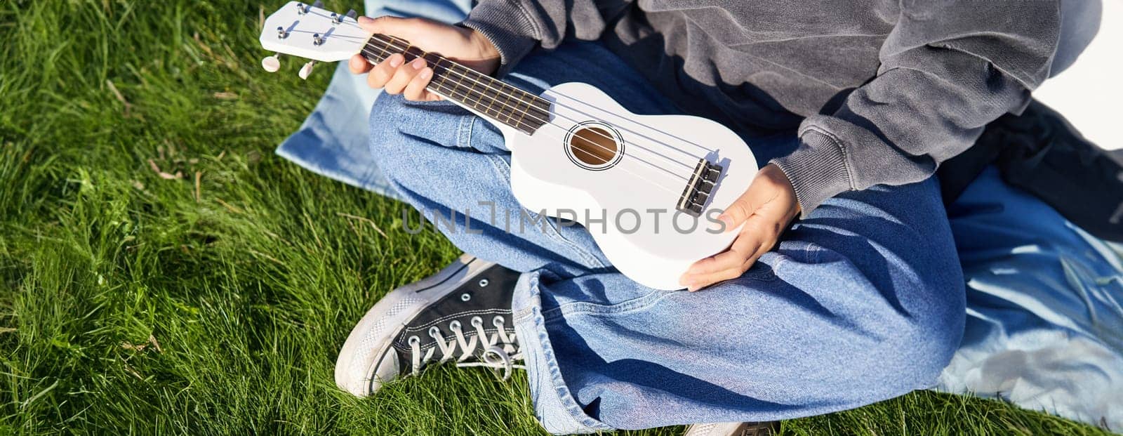 Music and instruments. Close up, female hands holding white ukulele, musician sitting on grass outdoors and playing.