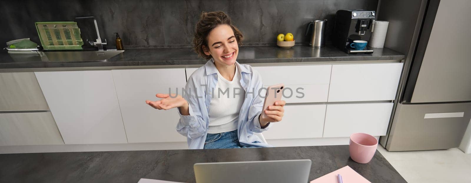 Chatty brunette woman sits at home, video chats with friend, waves hand at camera, holds smartphone and uses laptop.