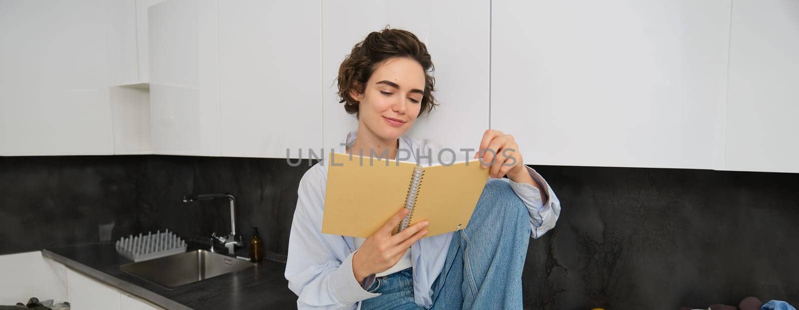 Portrait of young modern woman sitting in kitchen, flipping pages, reading journal book, smiling happily. People and lifestyle concept