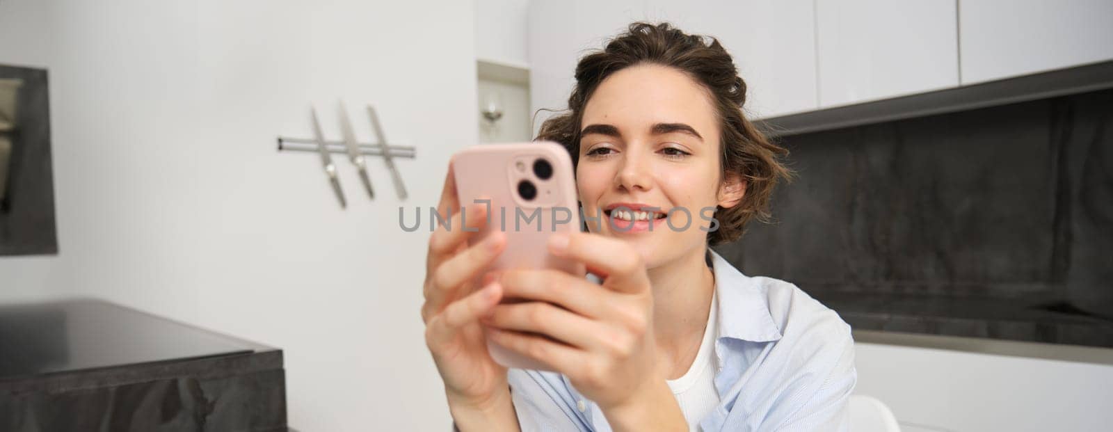 Image of young woman looking at her smartphone, reading message, sitting at home and making online purchase, paying for utilities on mobile app.