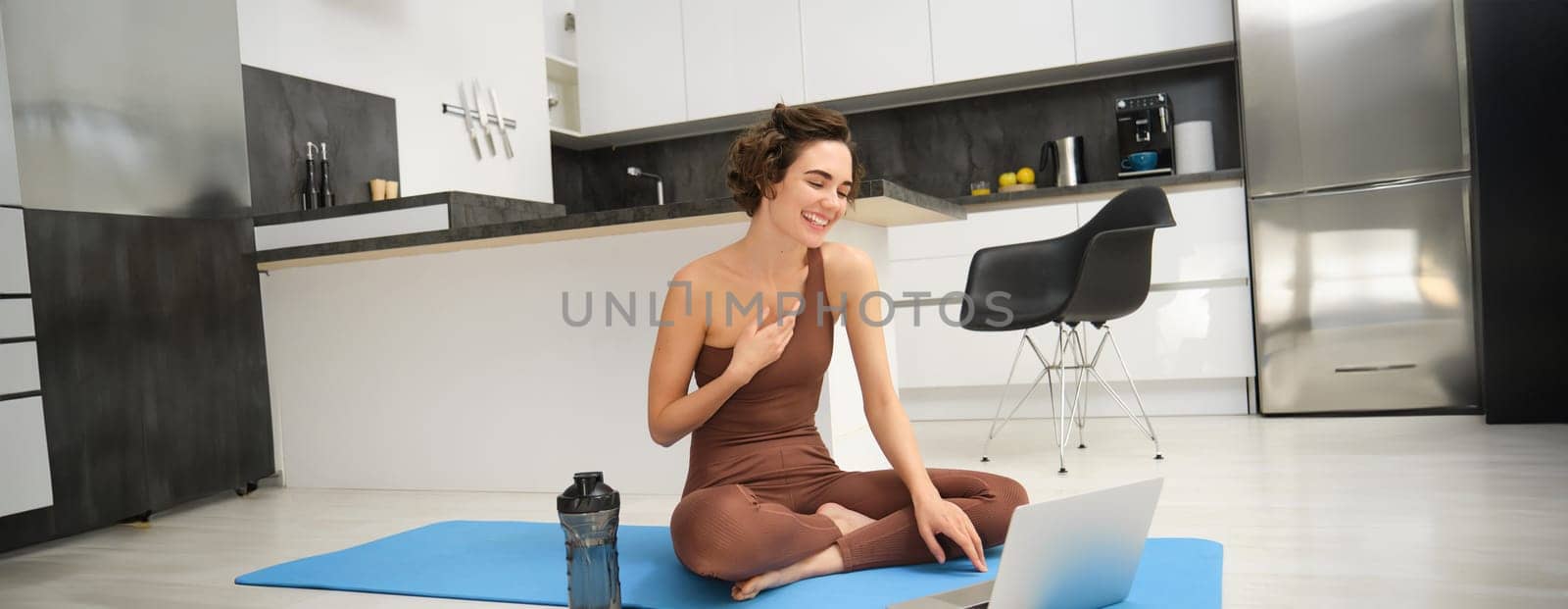 Sport and women. Young fitness instructor, yoga girl sits on rubber mat in her kitchen, waves hand and says hello at laptop, has online video gym class from home.