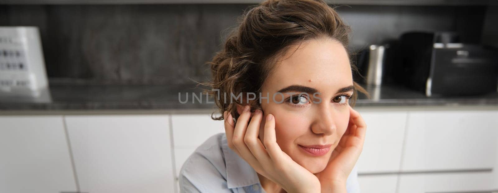 Close up portrait of young woman, 25 years old, sitting in her kitchen alone, express candid happiness, smiles, leans head on hands.