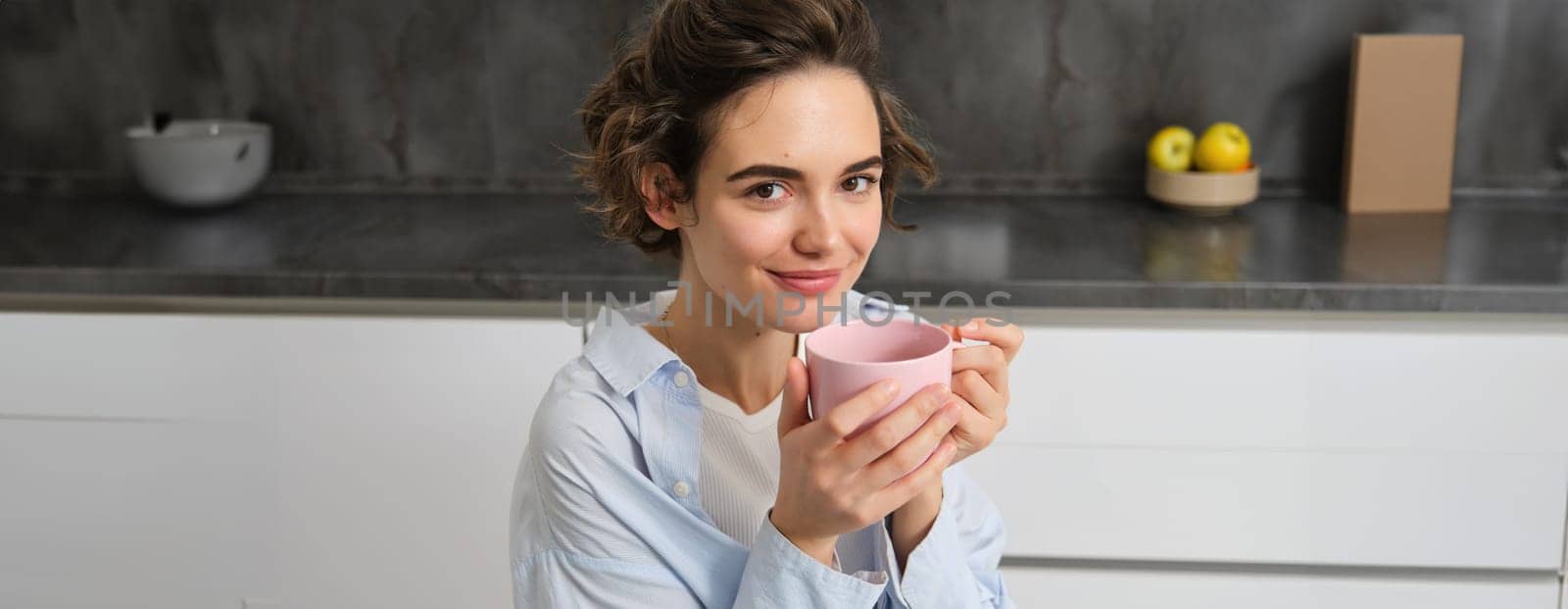Happy mornings. Portrait of happy brunette woman, drinks cup of coffee in her kitchen and smiling, cozy and warm start of the day with cuppa.