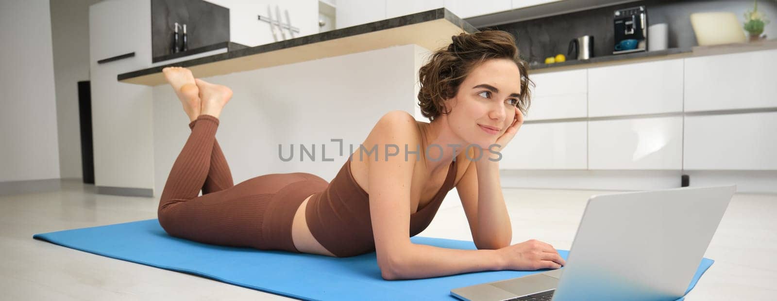 Portrait of woman athlete, girl workout at home, watches fitness videos on laptop, lying on rubber mat and doing indoor yoga session. Sport and wellbeing concept