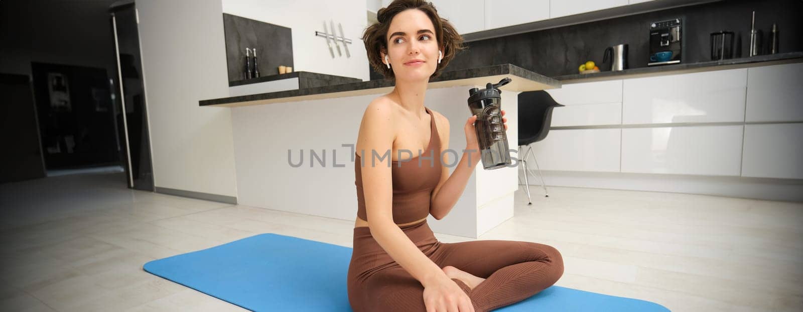 Portrait of fitness woman, young sportswoman at home, drinks water from bottle after workout, training exercises, takes a break after pilates, yoga training, stays hydrated.